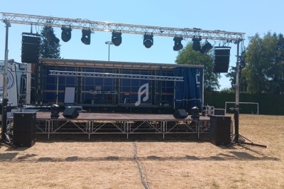 Scène extérieure avec structure métallique, éclairages suspendus et équipements audio sur un terrain herbeux.