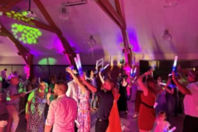 Des personnes dansent sous des lumières colorées dans une salle avec une charpente en bois, tenant des bâtons lumineux.