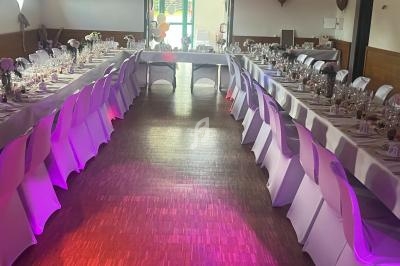 Salle décorée pour un événement avec des tables alignées, nappes blanches, chaises couvertes et ballons en arrière-plan.