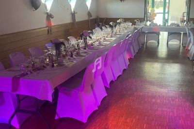 Salle décorée pour un événement avec deux longues tables dressées, nappes blanches et lumière tamisée.
