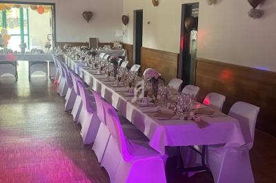 Salle décorée pour un événement avec une longue table dressée, nappes blanches, chaises couvertes et décorations murales.