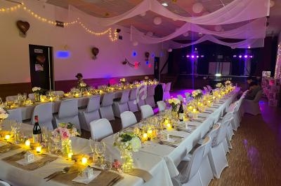 Salle décorée pour une réception avec longues tables dressées, bougies allumées et guirlandes lumineuses suspendues.