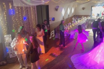 Des invités dansent et discutent dans une salle décorée pour un mariage, éclairée par des guirlandes lumineuses et des spots…