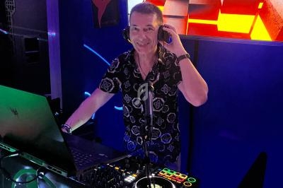 Un DJ souriant mixe de la musique dans un espace sombre, éclairé par des lumières colorées et un écran lumineux.