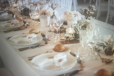 Table élégamment dressée avec nappes blanches, fleurs, verres, couverts et petits pains disposés pour un repas.