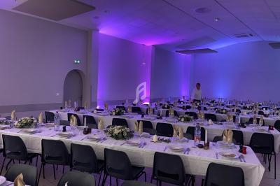 Salle de réception avec tables dressées, nappes blanches, décorations florales et éclairage violet.