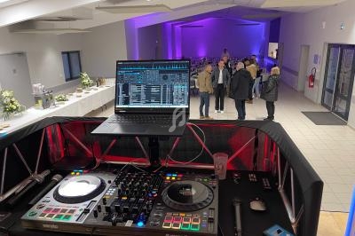 Poste de DJ avec platines et ordinateur en premier plan, salle de réception éclairée en arrière-plan avec invités.