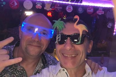 Deux hommes souriants portant des lunettes fantaisie dans un environnement festif et lumineux.