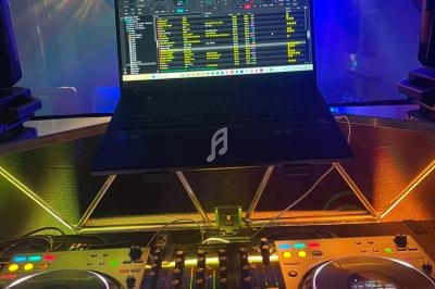 Poste de DJ avec platines, table de mixage et ordinateur affichant un logiciel de mixage, éclairé par des lumières colorées.