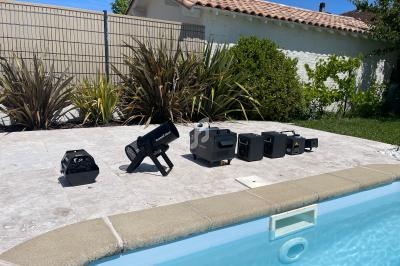 Équipements électroniques alignés sur une terrasse ensoleillée près d'une piscine, entourée de végétation et d'une maison.