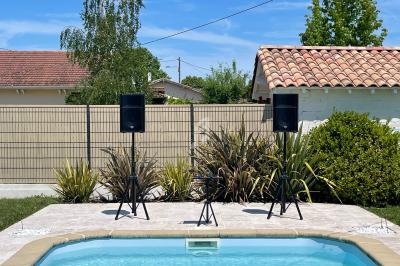 Deux enceintes sur trépieds installées près d'une piscine dans un jardin avec végétation et ciel dégagé.