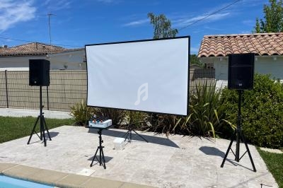 Écran de projection installé en extérieur, entouré de deux enceintes sur pieds, près d'une piscine et d'une maison.