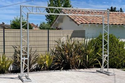 Structure métallique rectangulaire montée sur une terrasse carrelée, avec des plantes et une maison en arrière-plan.