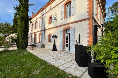 Façade d'une maison en briques et crépi avec volets bleus, terrasse pavée et jardin verdoyant.