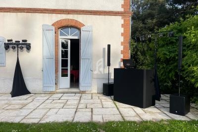 Matériel de sonorisation et éclairage installé sur une terrasse devant une maison avec volets bleus.