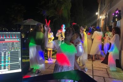 Groupe de personnes en tenue blanche dans une fête nocturne avec éclairages colorés, vue partielle d'une table de mixage.