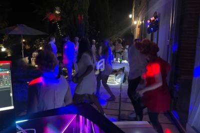 Des personnes dansent en soirée en plein air, éclairées par des lumières colorées, avec une table et des arbres en arrière…