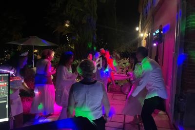 Des personnes dansent en soirée en extérieur, éclairées par des lumières colorées, près d'un bâtiment et d'arbres.