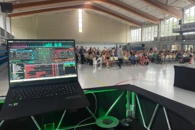 Vue d'un ordinateur portable avec un logiciel de mixage audio, face à un public assis dans une salle polyvalente lumineuse.