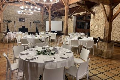 Salle de réception avec tables rondes dressées, chaises blanches, poutres apparentes et murs en pierre.