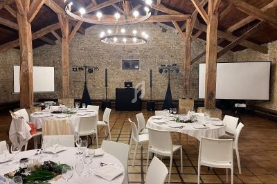 Salle de réception avec tables rondes dressées, chaises blanches, murs en pierre et poutres apparentes en bois.