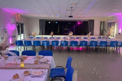 Salle décorée pour un événement avec longues tables dressées, chaises bleues et éclairage coloré sur une scène au fond.