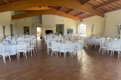 Salle de réception lumineuse avec tables rondes dressées, chaises blanches et poutres apparentes au plafond.