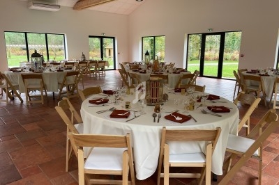 Salle de réception lumineuse avec tables rondes dressées, nappes blanches et serviettes rouges, vue sur un jardin.