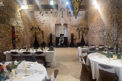 Salle en pierre ancienne décorée pour un événement, avec tables dressées, chaises couvertes et éclairage tamisé.