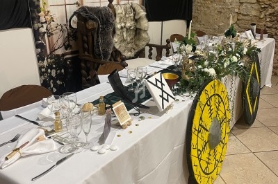 Table décorée dans un style médiéval avec vaisselle, boucliers ornés et éléments floraux, dans une salle en pierre.