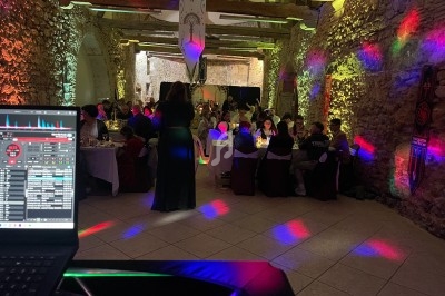 Salle en pierre éclairée par des lumières colorées, avec des convives assis à des tables et un DJ en arrière-plan.