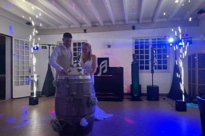 Un couple en tenue de mariage coupe un gâteau posé sur un tonneau, dans une salle décorée avec des lumières festives.