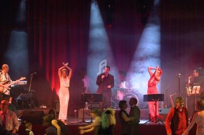 Des musiciens et chanteurs sur scène avec des danseurs au premier plan dans une ambiance lumineuse et festive.