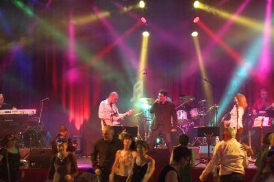 Groupe de musiciens jouant sur scène avec des éclairages colorés, devant un public dansant.