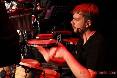 Un musicien joue des percussions sur scène, éclairé par une lumière rouge.