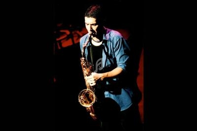 Un musicien joue du saxophone sur une scène sombre, éclairé par une lumière chaude.