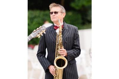 Un homme en costume rayé joue du saxophone en extérieur, avec une guitare visible en arrière-plan.