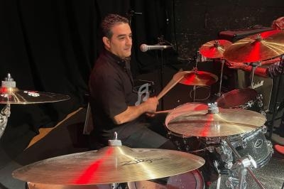 Un batteur joue sur une batterie argentée sur une scène sombre, avec un micro à proximité.