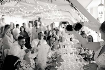 Des invités applaudissent tandis qu'une mariée remplit une pyramide de coupes avec du champagne lors d'une réception.