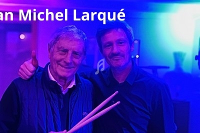 Deux hommes posant ensemble dans une ambiance lumineuse bleue et violette, l'un tenant des baguettes de batterie.