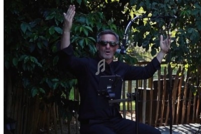 Un homme en lunettes de soleil chante ou parle au micro, bras levés, devant un fond de végétation et une clôture en bois.