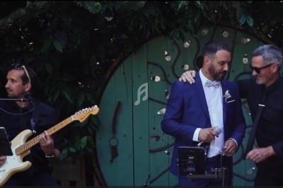 Un homme en costume bleu et un autre en noir se tiennent près d'un micro, avec un guitariste jouant à gauche.