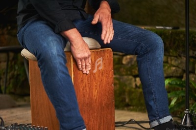Musicien jouant du cajón en bois, ses mains frappant le devant de l'instrument dans un cadre extérieur.