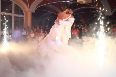 Un couple en tenue de mariage danse entouré de fumée et d'étincelles, devant un public enthousiaste.