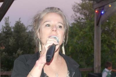 Femme tenant un microphone et chantant en extérieur sous une pergola éclairée, avec un fond de végétation.