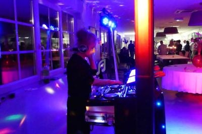 Une DJ anime une soirée dans une salle éclairée par des lumières colorées, avec des invités en arrière-plan.