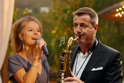 Une femme chante au micro tandis qu'un homme joue du saxophone en extérieur, entourés de verdure.