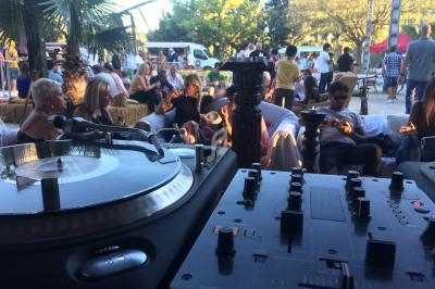 Platines de DJ au premier plan avec des personnes assises en arrière-plan dans un espace extérieur animé.
