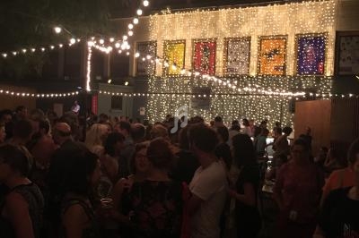 Foule rassemblée de nuit dans une cour éclairée par des guirlandes lumineuses, avec des affiches colorées en arrière-plan.