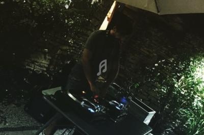 Un DJ mixe de la musique sur une table de mixage en extérieur, sous un parasol, entouré de végétation.